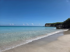 Ein Strand mit klarem türkisblauem Wasser, sandigem Ufer und einer felsigen Klippe mit Vegetation.