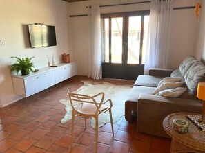 Un saló amb un sofà, una cadira, una televisió penjada a la paret, una finestra amb cortines i una taula petita amb una làmpada.