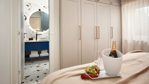 Una camera d'hotel con un letto, un comodino con una bottiglia di champagne, un bicchiere e degli acini di uva. Si intravede un bagno attraverso una porta aperta.