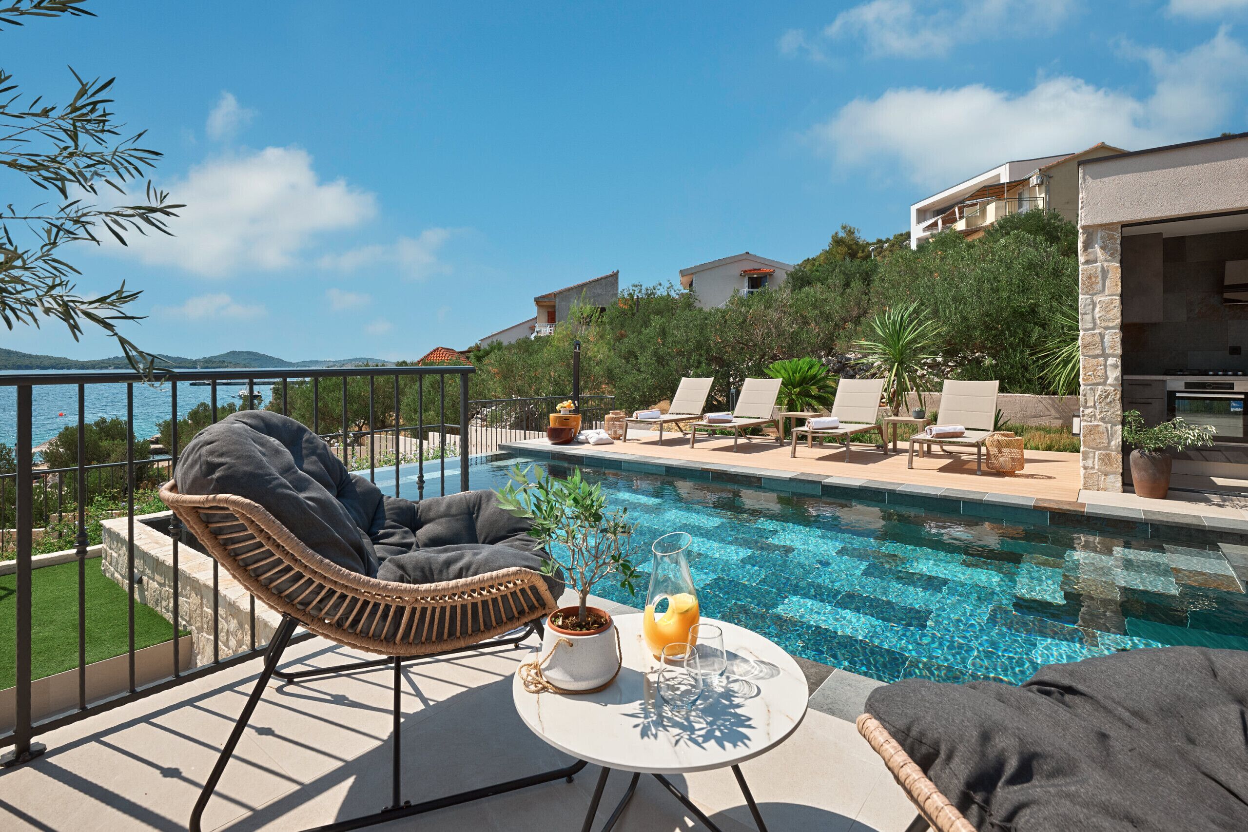 Un espace aménagé au bord de la piscine, avec des chaises longues, une table où sont posées des boissons, et une vue sur la mer et les bâtiments.