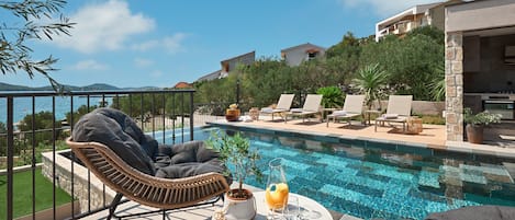 Zona junto a la piscina con sillas de descanso, una mesa con una bebida y vistas al mar y edificios.
