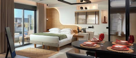 A modern hotel room with a bed, bedside table, and a dining area with red plates and glasses.