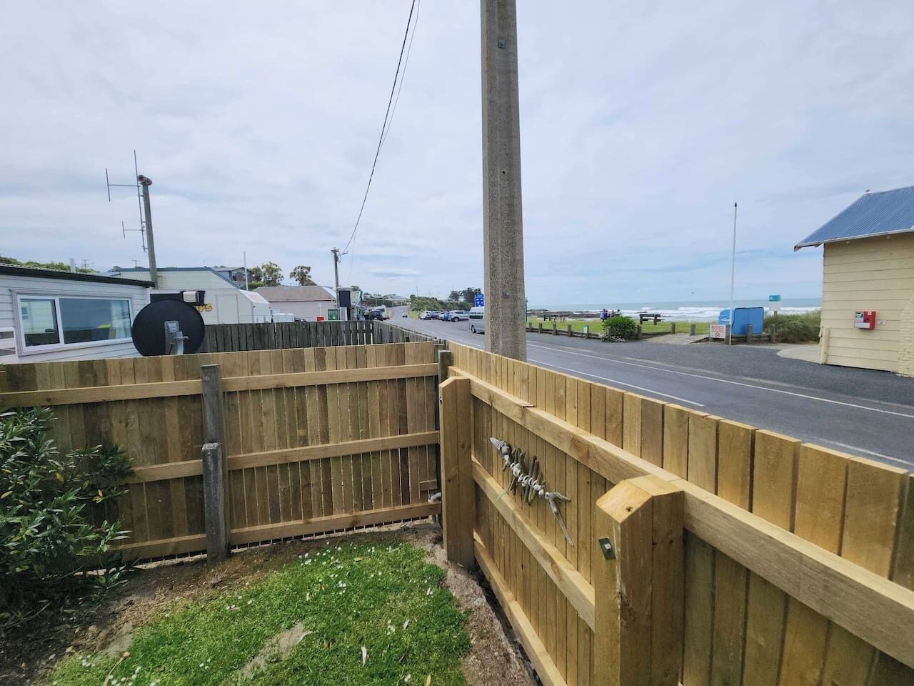 A wooden fence with a gate, a road, and a building with a blue roof.