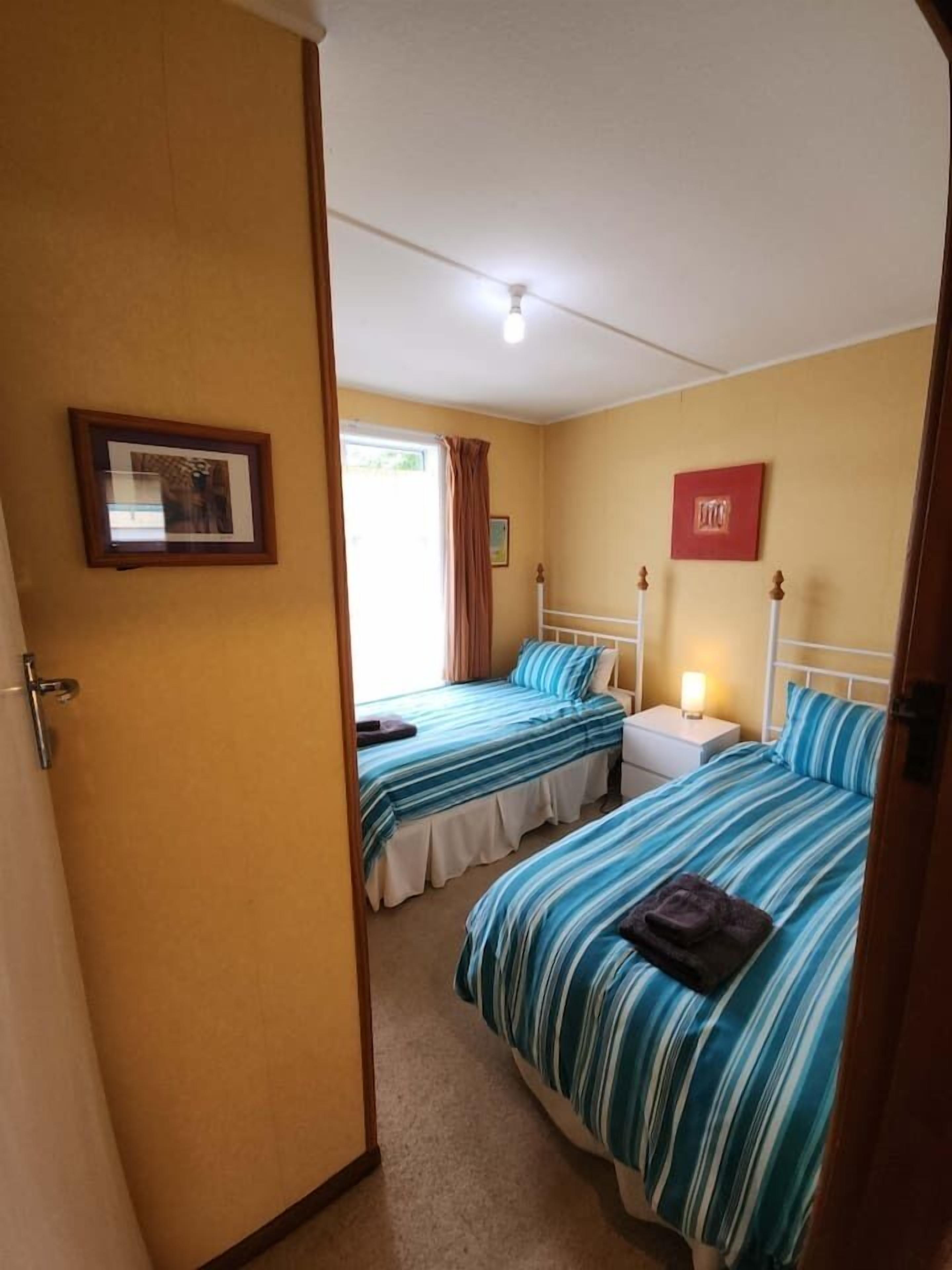 A room with two beds, a bedside table with a lamp, a framed picture on the wall, and a door.
