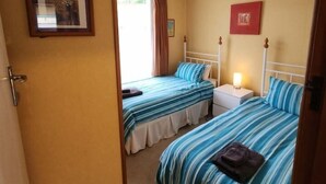 A room with two beds, a bedside table with a lamp, a framed picture on the wall, and a door. - Esplanade Escape – Classic Coastal Cottage (Kaka Point)