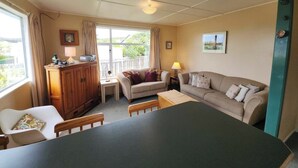 A room with a dining table, two sofas, a wooden cabinet, and a window with curtains. - Esplanade Escape – Classic Coastal Cottage (Kaka Point)