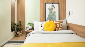 A modern bedroom with a bed, a framed artwork, a bedside table with a plant, and a wall-mounted light. - Unite Students - Emily Bowes - London (London)