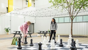 A chessboard set up outdoors with two people playing, a tree, and picnic tables. - Unite Students - Emily Bowes - London (London)
