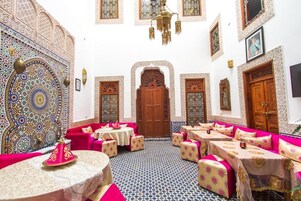 Um interior tradicional marroquino com azulejos ricamente decorados, uma porta de madeira central e áreas de estar com almofadas estampadas.