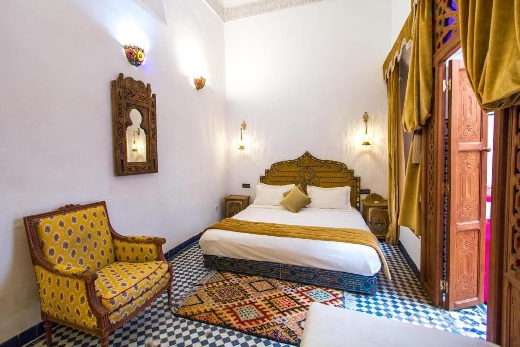 A bedroom with a bed, a chair, a patterned rug, and a wall-mounted light fixture.