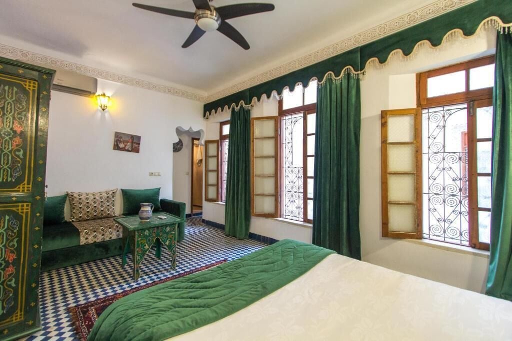Una habitación con cama, sofá, una mesita, ventilador de techo y ventanas con diseño.