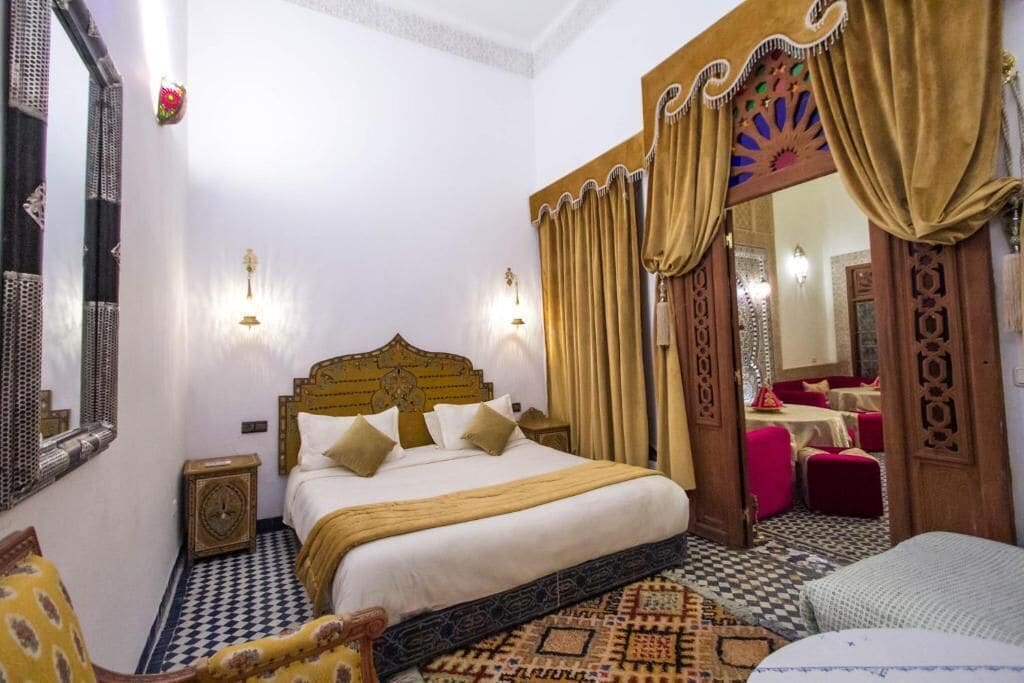 Ein Zimmer mit einem Bett, einem Spiegel, einem Nachttisch und einem gemusterten Teppich.