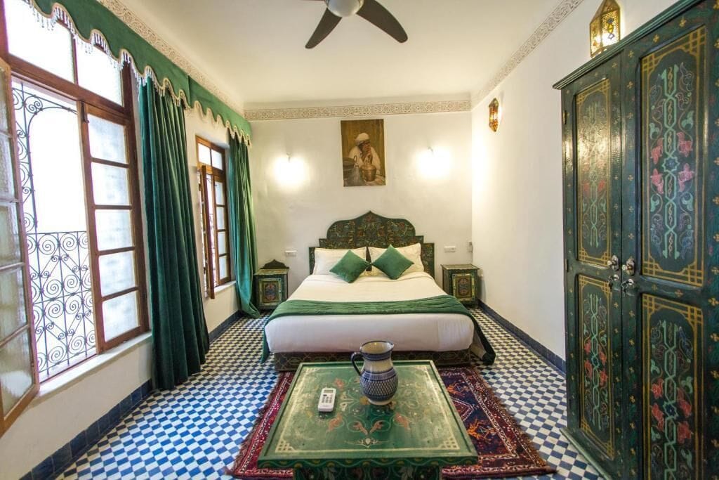 A room with a bed, a table with a pitcher, patterned curtains, a checkered floor, and a decorative cabinet.