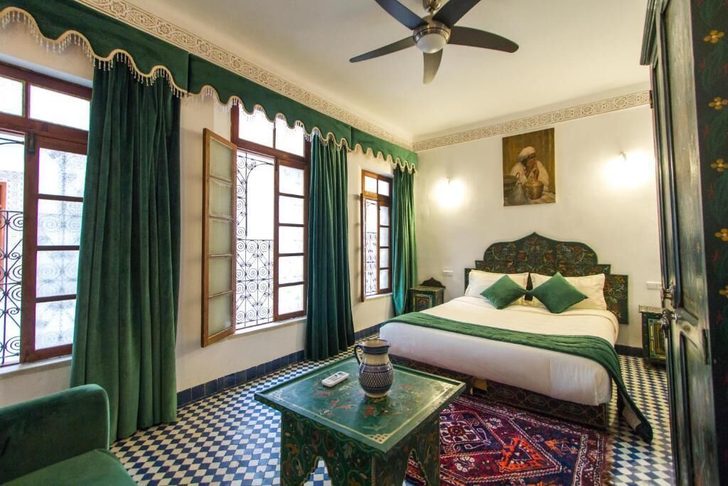 A room with a bed, a ceiling fan, a table with a pitcher, patterned curtains, and a checkered floor.