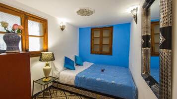 A bedroom with a blue wall, a wooden door, a bed with blue bedding, a patterned rug, a bedside lamp, and a vase with flowers.