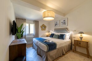 Quarto de hotel com cama, mesas de cabeceira, televisão, planta e obras de arte nas paredes.
