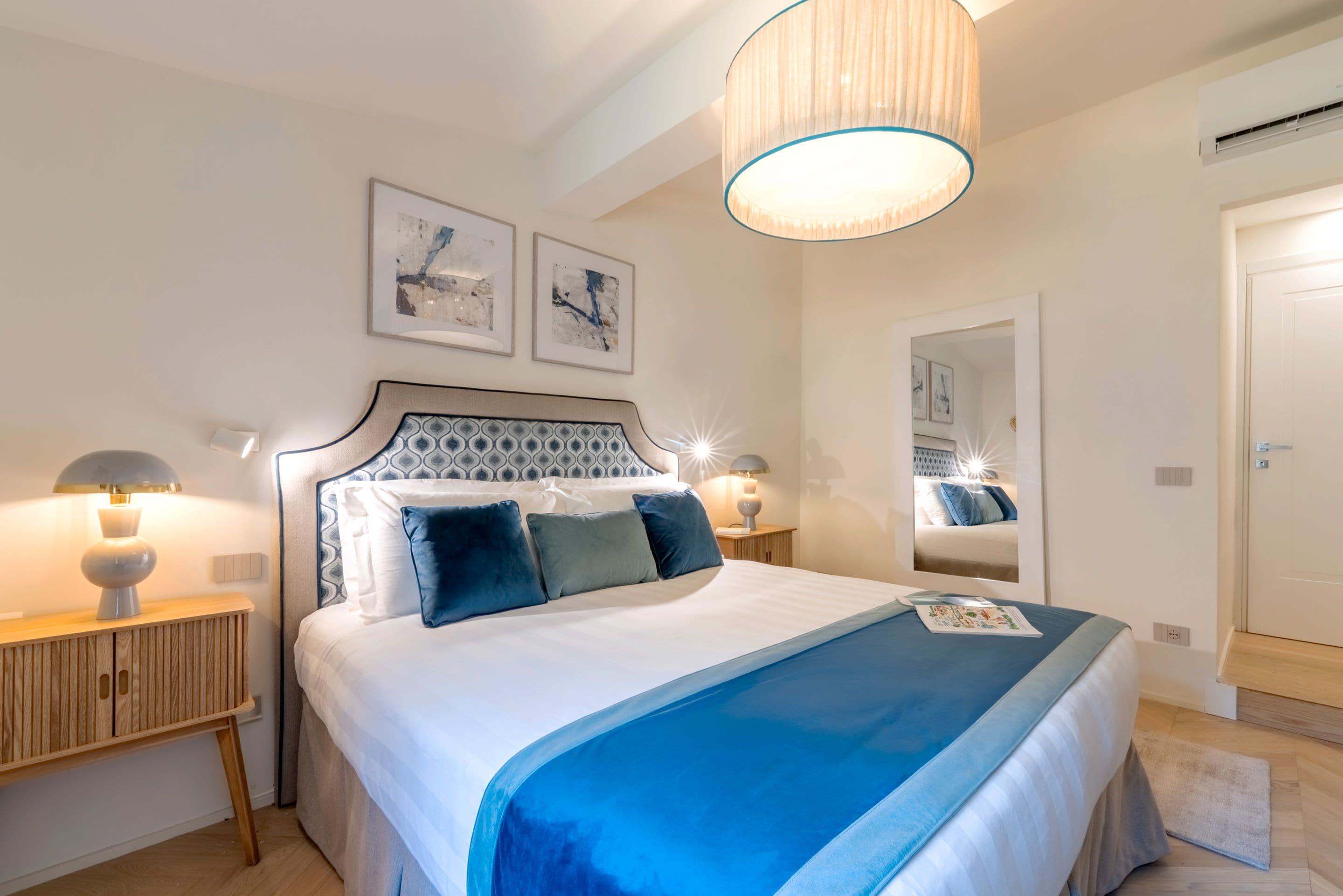 A bedroom with a bed, bedside lamps, a mirror, and artwork on the wall.