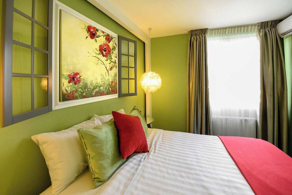 Ein Hotelzimmer mit Bett, Kissen, einem Gemälde, einer Lampe und einem Fenster mit Vorhängen.
