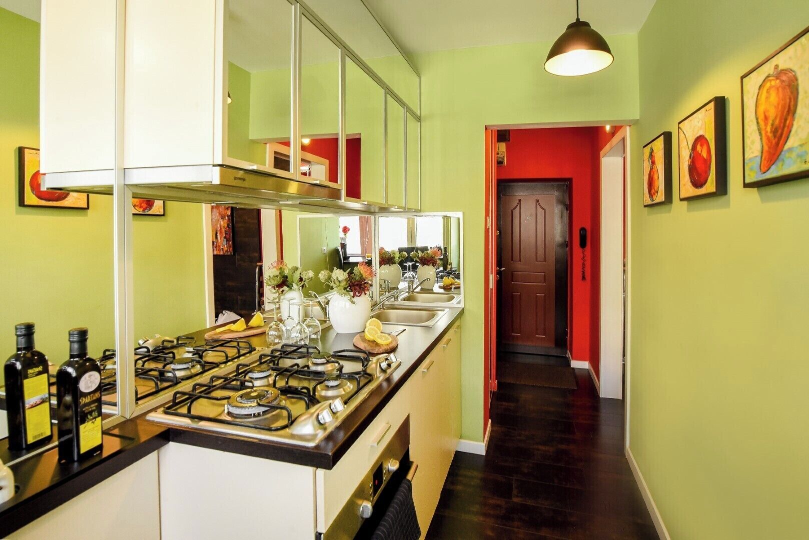 A kitchen with a gas stove, a sink, and a mirror. There is a hallway with a red door and a staircase.