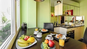 Ontbijt met fruit, koffie en sap op een tafel in een kamer met raam, stoel en een keukenhoek.