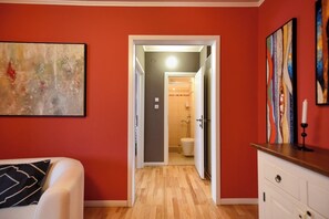 Ein Zimmer mit Holzboden, einem Bett mit Kopfkissen in Schwarzweiß, einer weißen Kommode und einem durch eine offene Tür einsehbaren Badezimmer.