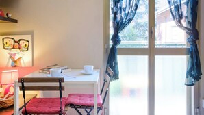 Een kamer met een tafel, stoelen, een raam met gordijnen, een lamp en een wand met een ingelijste foto.