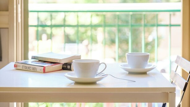 Een tafel met twee witte kopjes en schotels, een boek en een raam met een groene balustrade.