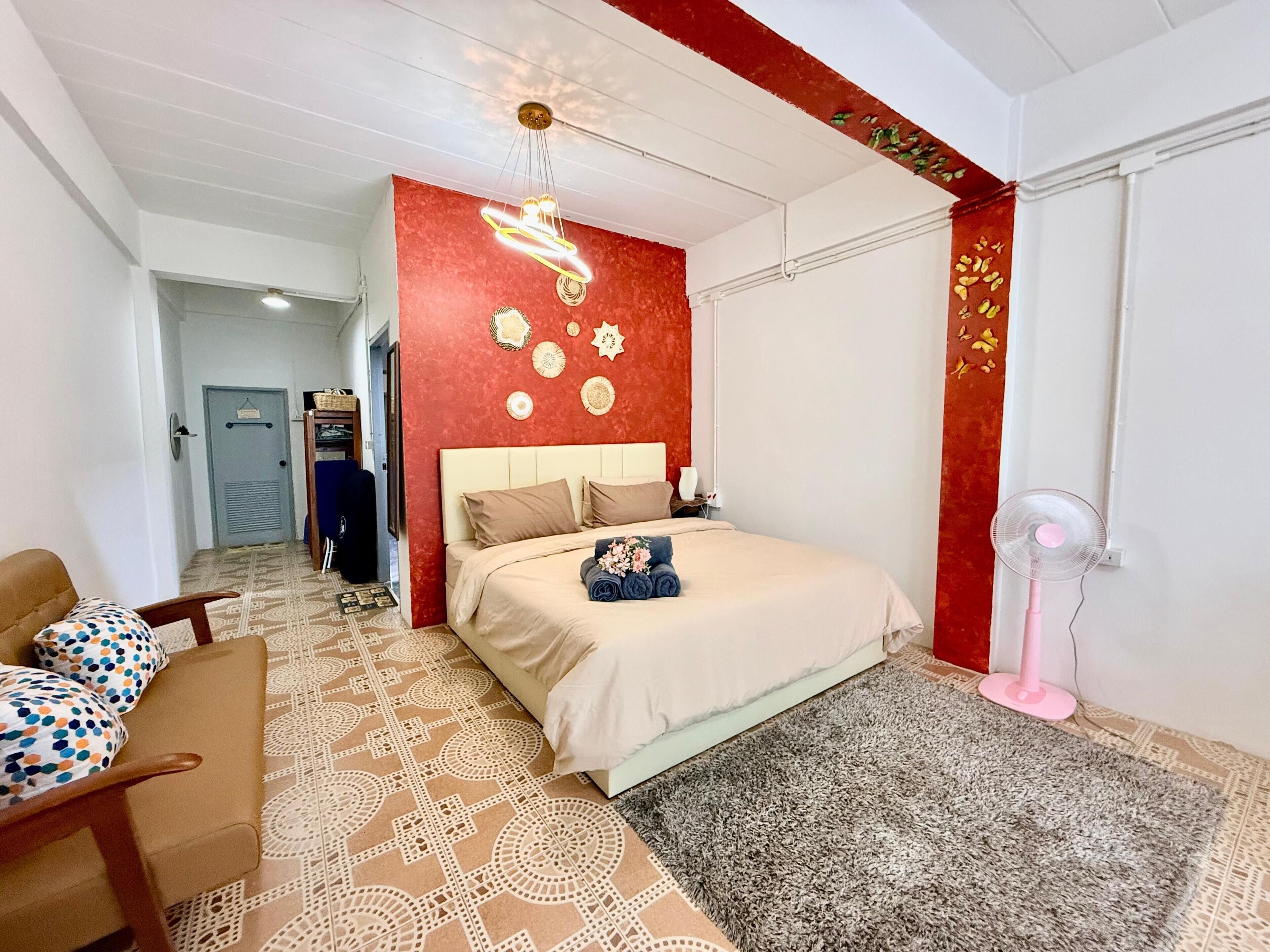 Una habitación de hotel con una cama, una silla, un escritorio, una lámpara de escritorio, un reloj de pared y un ventilador de techo.