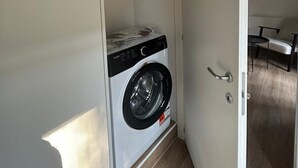 Quarto compacto com máquina de lavar e secar embutidas, porta com puxador e um pequeno espaço de estar com cadeira e mesa.