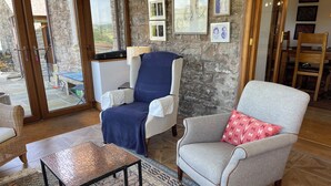 Una habitación con pared de piedra, un ventanal grande, un sillón azul, un sillón gris con un cojín rojo y una alfombra estampada.
