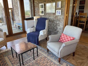 A room with a stone wall, a large window, a blue armchair, a grey armchair with a red pillow, and a patterned rug.