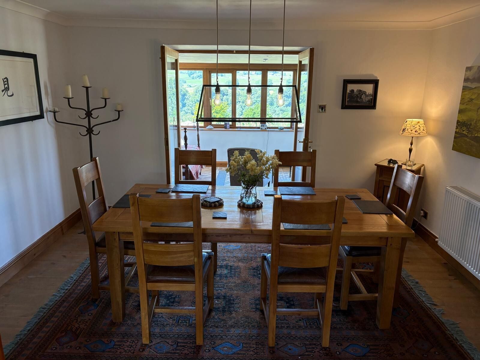 Une salle à manger avec une table en bois, six chaises, un lustre, un vase rempli de fleurs et une vue par la fenêtre.