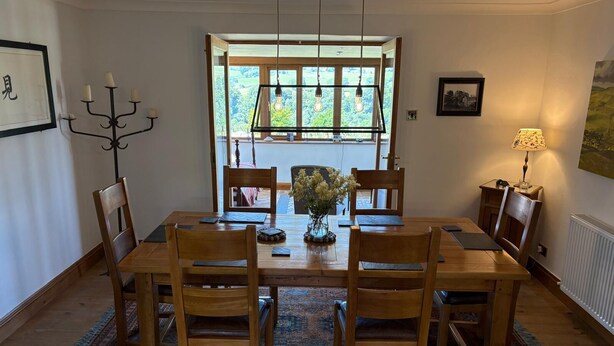 Un comedor con una mesa de madera, seis sillas, una lámpara de araña, un jarrón con flores y vistas al exterior por una ventana.