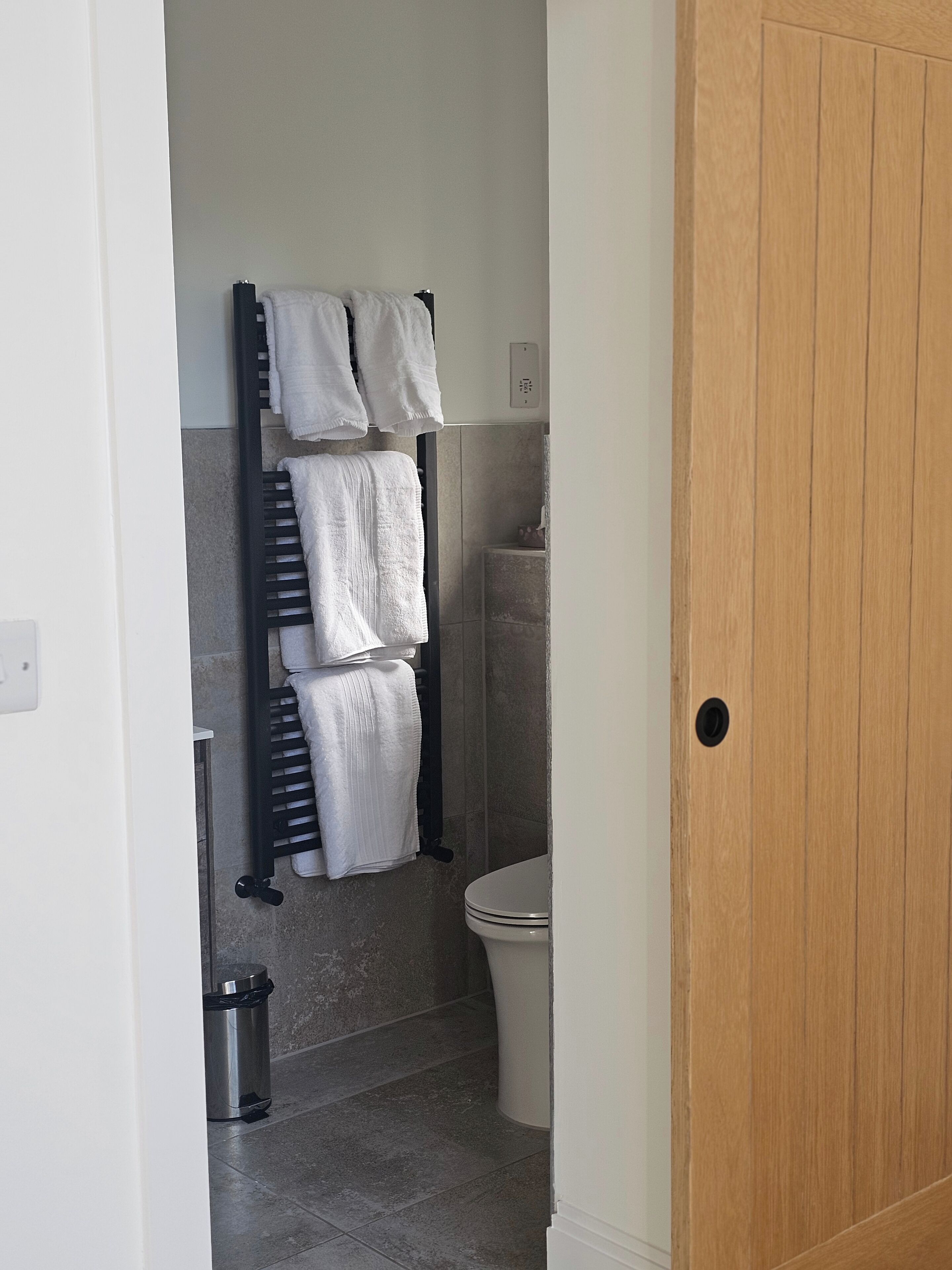 A bathroom with a toilet, a towel rack with multiple towels, a trash bin, and a wooden door.