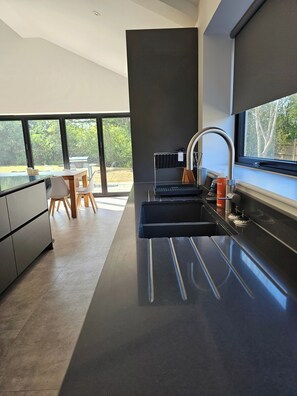 A modern kitchen with a large window, a dining area with a table and chairs, and a view of greenery outside. - Luxury Kent Bungalow Retreat Sleeps 12 (Tonbridge)