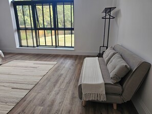 Une pièce comprenant un canapé, une lampe sur pied, une fenêtre avec des stores et donnant sur une terrasse en bois.