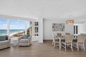 A modern living room with a large window overlooking a beach, a dining area with a chandelier, and a view of a building across the street. - Luxury 3BR Beachfront Condo - Wrap Balcony, Endless Gulf Views! Pool & Beach! (Miramar Beach)