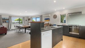 A modern kitchen with a dishwasher, stove, and dining area with a table and chairs. - Wanaka 'Mount Iron Edge' with Mountain Views (Wanaka)