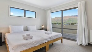 A hotel room with a large bed, two bedside tables, a nightstand, and a view of the mountains through a large window. - Wanaka 'Mount Iron Edge' with Mountain Views (Wanaka)
