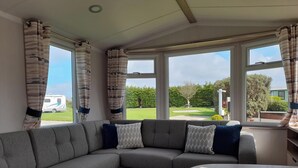 Ein modernes Wohnzimmer mit einem grauen Ecksofa, einem Holztisch und großen Fenstern mit Blick auf einen Garten und einen abgestellten Wohnwagen.