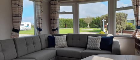 A modern living room with a grey sectional sofa, a wooden coffee table, and large windows offering a view of a garden and a parked caravan.