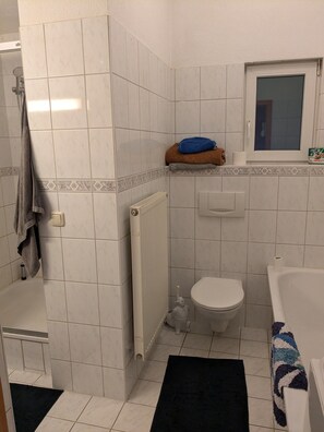 Een badkamer met witte tegels, een toilet, een badkuip, een handdoekhouder en een raam.