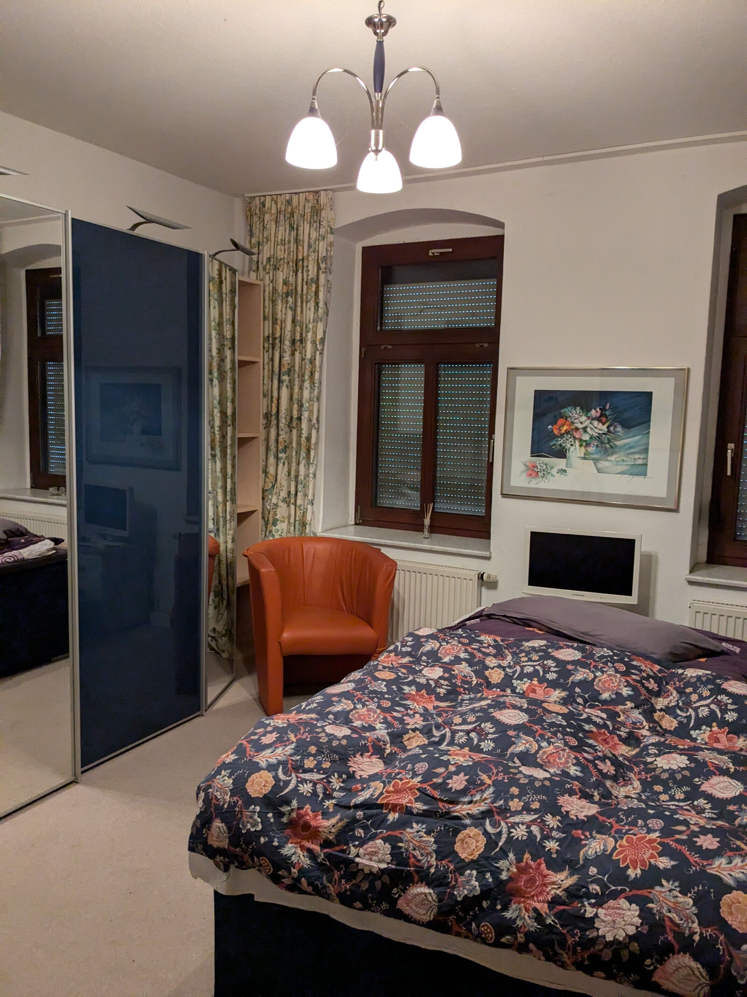 Ein Schlafzimmer mit einem Bett, einem Sessel, einem Fernseher, einem Fenster mit Jalousien, einer Heizung und einer Deckenleuchte.