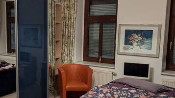 Ein Schlafzimmer mit einem Bett, einem Sessel, einem Fernseher, einem Fenster mit Jalousien, einer Heizung und einer Deckenleuchte.
