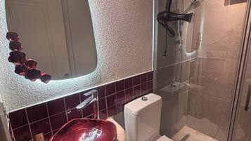 A bathroom with a glass shower enclosure, a white toilet, and a red sink with a decorative mirror above it.