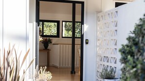 Une terrasse en bois mène à un bâtiment blanc doté d’une porte à cadre noir, d’une plante en pot et d’un mur en treillis blanc.