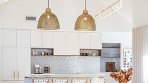Une cuisine moderne avec des meubles blancs, un îlot central, des luminaires suspendus et un coin repas avec des chaises et une table en bois.