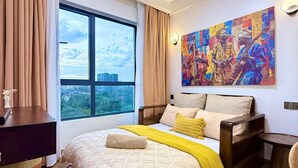 A bedroom with a large window, a bed with a patterned bedspread, a bedside table, a nightstand, and a wall-mounted painting. - Capital Rise View Palace Apartment (Nairobi)