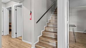 Una escalera con escalones de madera y barandilla metálica, paredes blancas y un extintor de incendios montado en la pared.