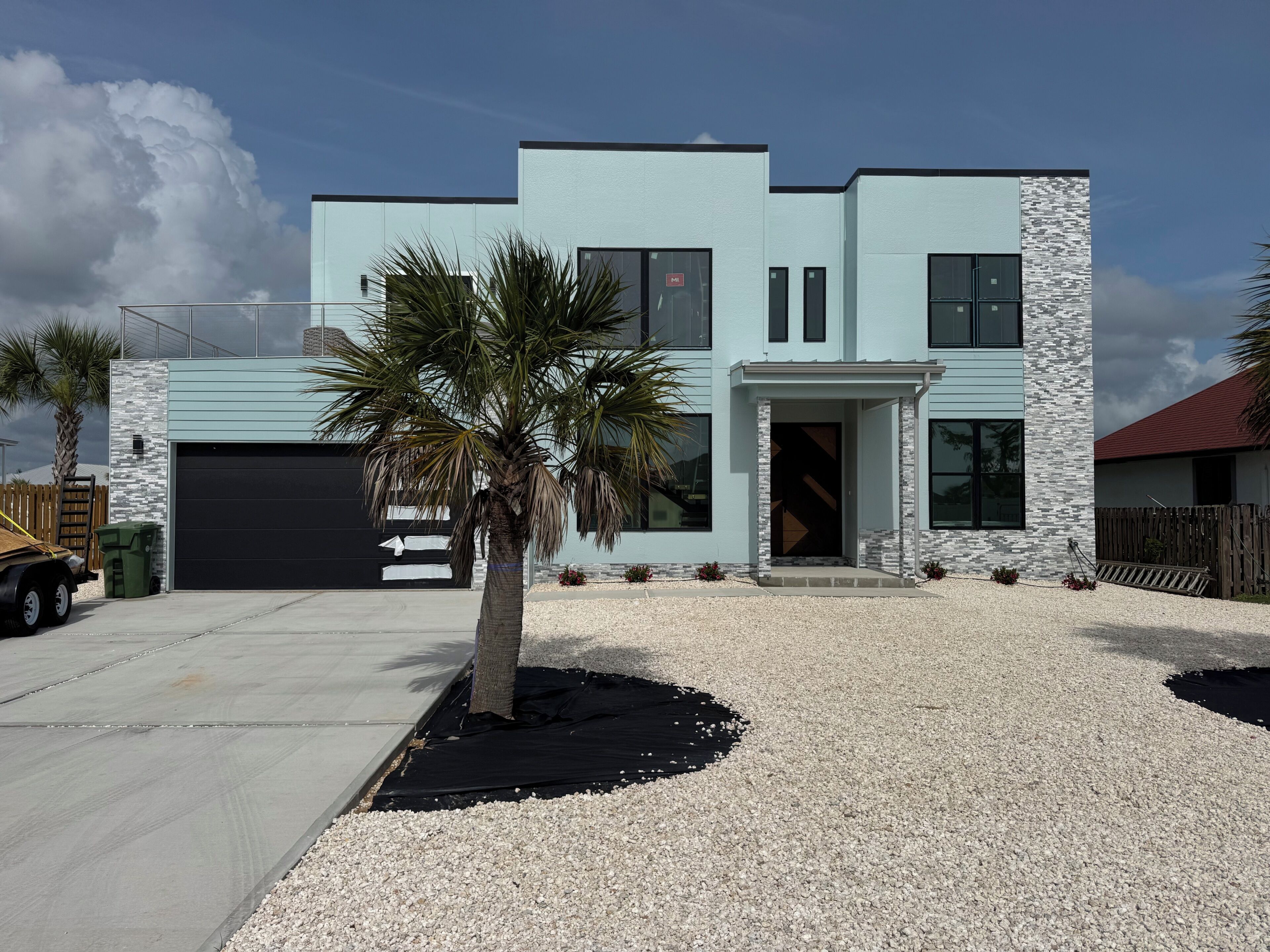 A modern two-story house with a garage, a palm tree, and a gravel driveway.